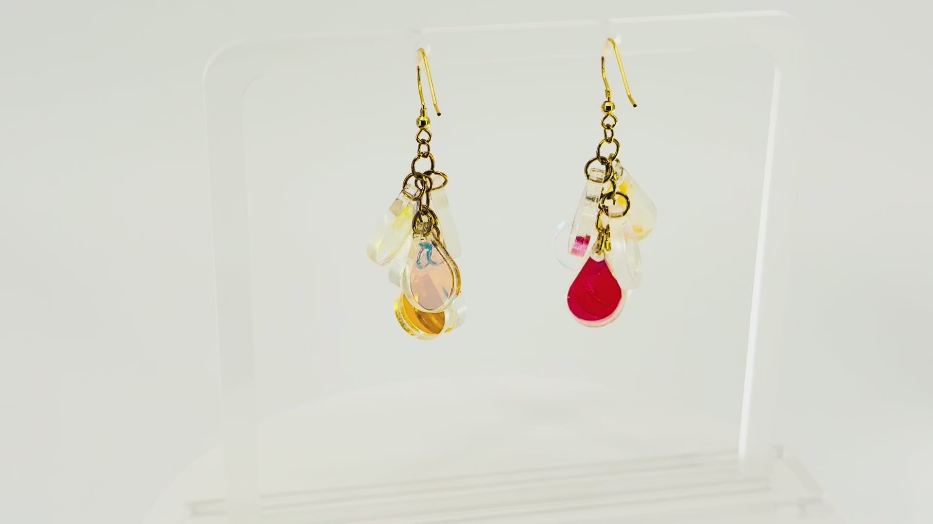 A pair of dangling earrings displayed on a clear acrylic stand. Each earring has a gold fishhook ear wire with a cluster of dangling iridescent teardrop shaped charms. The stand is being rotated for the video.