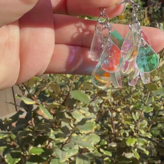 A hand holds a pair of dangling earrings displayed in the sunshine in front of some plants. Each earring has a silver fishhook ear wire with a cluster of dangling iridescent teardrop shaped charms.