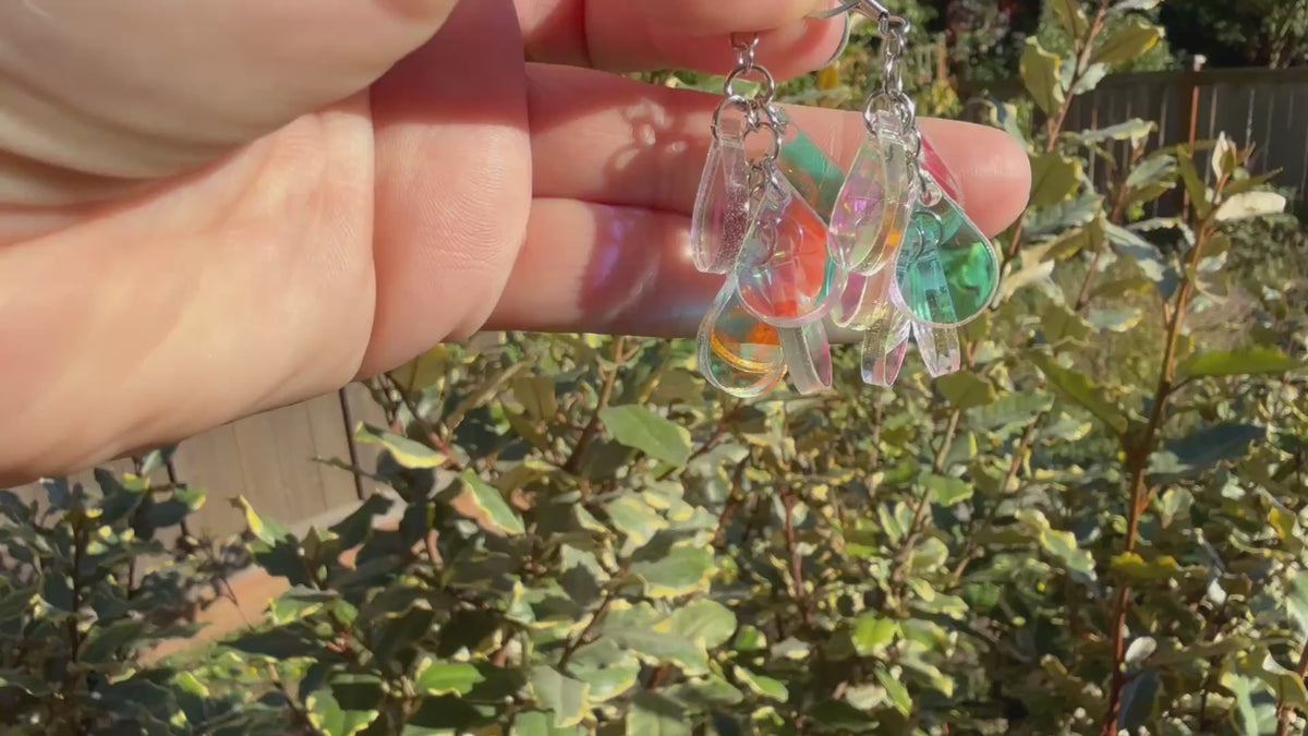 A hand holds a pair of dangling earrings displayed in the sunshine in front of some plants. Each earring has a silver fishhook ear wire with a cluster of dangling iridescent teardrop shaped charms.