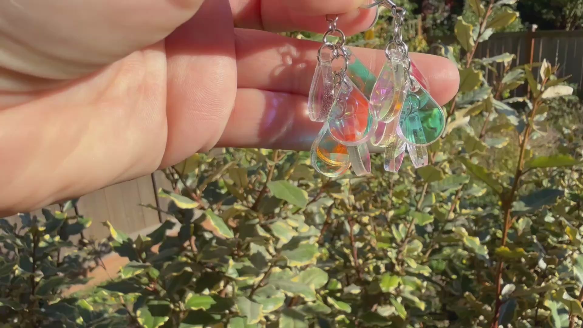 A hand holds a pair of dangling earrings displayed in the sunshine in front of some plants. Each earring has a silver fishhook ear wire with a cluster of dangling iridescent teardrop shaped charms.